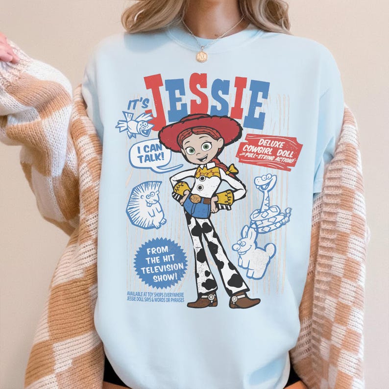 May include: Light blue t-shirt featuring a graphic of Jessie from the Toy Story franchise. The design includes the text "JESSIE", "I CAN TALK!", and "DELUXE COWGIRL DOLL". Additional graphics of a snake, a rabbit, and a hedgehog are also present.