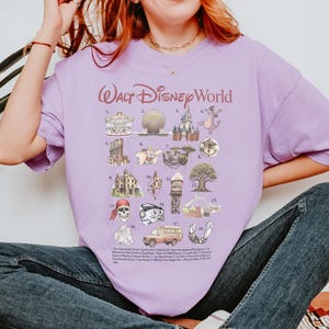 K&ouml;nnte beinhalten: Helllila T-Shirt mit der Aufschrift "Walt Disney World" und 18 farbigen Illustrationen von Parkattraktionen. Das T-Shirt enth&auml;lt eine Liste der Attraktionen unter den Illustrationen.