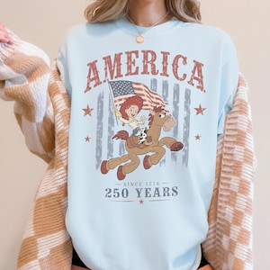 May include: A light blue crewneck sweatshirt featuring a graphic of a cowgirl riding a brown horse while holding an American flag. The text reads AMERICA, SINCE 1776, and 250 YEARS, accompanied by red stars and a faded vertical striped background design.