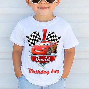 May include: White t-shirt featuring a red race car graphic with the number 1, checkered flags, and the name "David" in a red banner. The text "Birthday boy" is also displayed in red. A fun, themed shirt for a child's birthday.
