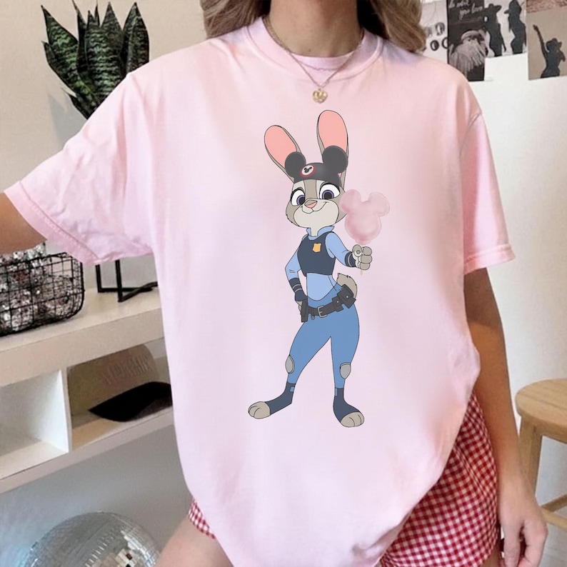 Puede incluir: Camiseta rosa claro con una ilustraci&oacute;n de dibujos animados de Judy Hopps de Zootopia. Judy, con su uniforme de polic&iacute;a, sostiene un algod&oacute;n de az&uacute;car rosa. La camiseta es de cuello redondo.
