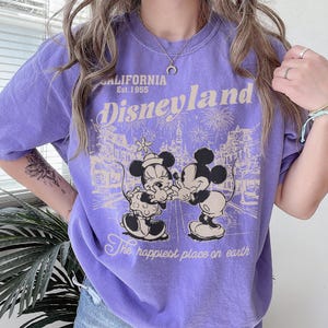 May include: A purple t-shirt with a Disneyland graphic. The graphic features Mickey and Minnie Mouse, fireworks, and the text "CALIFORNIA Est. 1955 Disneyland The happiest place on earth."