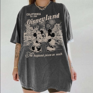 May include: Dark gray t-shirt featuring a Disneyland graphic. The design includes the text "CALIFORNIA Est. 1955 Disneyland" above an illustration of Mickey and Minnie Mouse. The phrase "The happiest place on earth" is below the illustration.