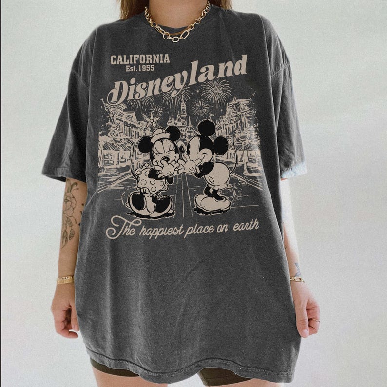 May include: Dark gray t-shirt featuring a Disneyland graphic. The design includes the text "CALIFORNIA Est. 1955 Disneyland" above an illustration of Mickey and Minnie Mouse. The phrase "The happiest place on earth" is below the illustration.