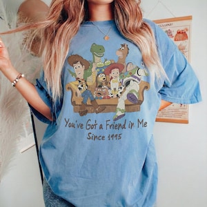 May include: A blue t-shirt featuring a graphic print of Toy Story characters sitting on a couch. The characters include Woody, Buzz Lightyear, Jessie, Rex, Slinky Dog, and Mr. Potato Head. Below the graphic, the text reads: You've Got a Friend in Me Since 1995.