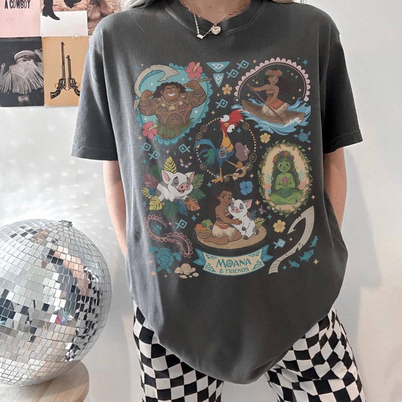 May include: Dark gray t-shirt featuring a colorful graphic of characters from the animated film "Moana." The design includes images of Maui, Moana, Pua, Hei Hei, and other elements. The words "Moana & Friends" are printed at the bottom.