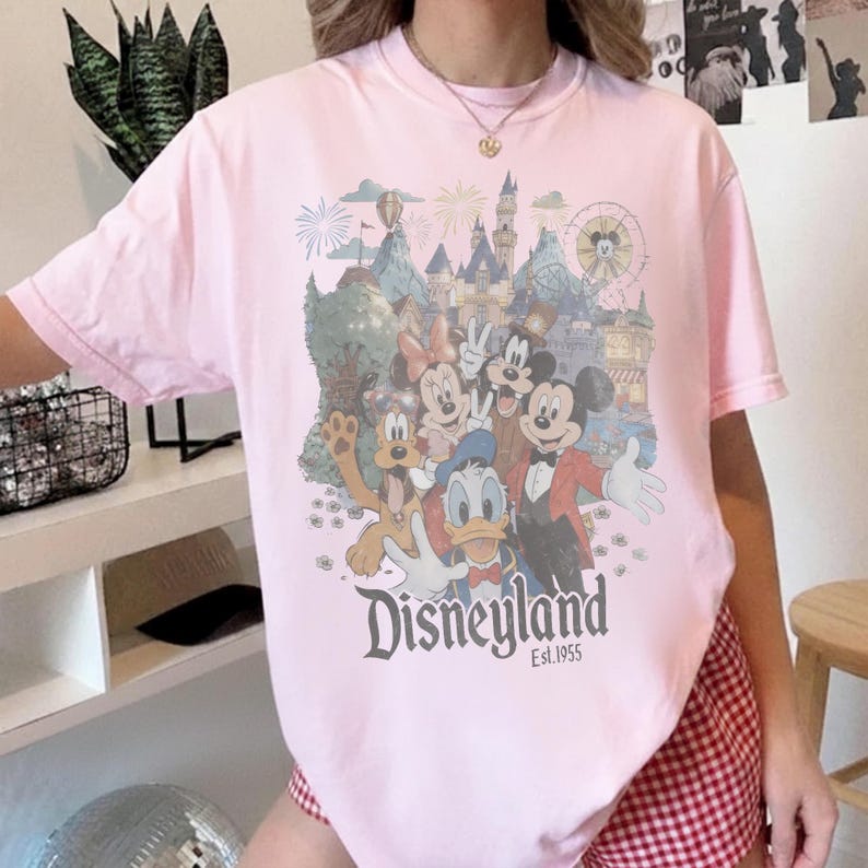 Puede incluir: Camiseta rosa claro con un gr&aacute;fico de Disneyland. El gr&aacute;fico presenta a Mickey Mouse, Minnie Mouse, Donald Duck, Goofy y un castillo. Tambi&eacute;n se ve el texto "Disneyland Est.1955".