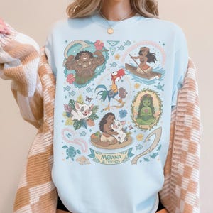 May include: Light blue sweatshirt featuring colorful illustrations of characters from the animated film "Moana." The design includes images of Maui, Moana, Pua, Hei Hei, and other characters, with the text "Moana & Friends" at the bottom.