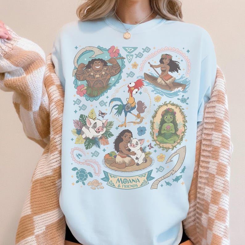 May include: Light blue sweatshirt featuring colorful illustrations of characters from the animated film "Moana." The design includes images of Maui, Moana, Pua, Hei Hei, and other characters, with the text "Moana & Friends" at the bottom.