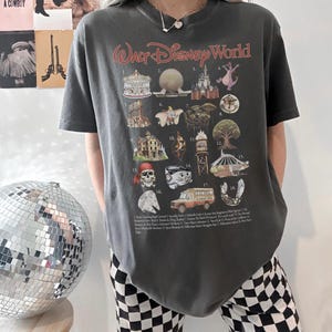 K&ouml;nnte beinhalten: Dunkelgraues T-Shirt mit einem Walt Disney World-Motiv, das verschiedene Parksymbole und den Text "Walt Disney World" in roter Schrift zeigt. Das T-Shirt hat einen Vintage-Look.