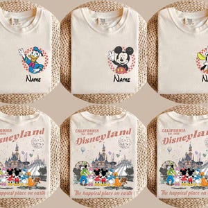 K&ouml;nnte beinhalten: Cremefarbene T-Shirts mit den Disney-Figuren Donald Duck, Mickey Mouse und Goofy, jeweils mit einem Namensfeld. Darunter zeigen drei Shirts eine Disneyland-Grafik mit dem Text "The happiest place on earth."