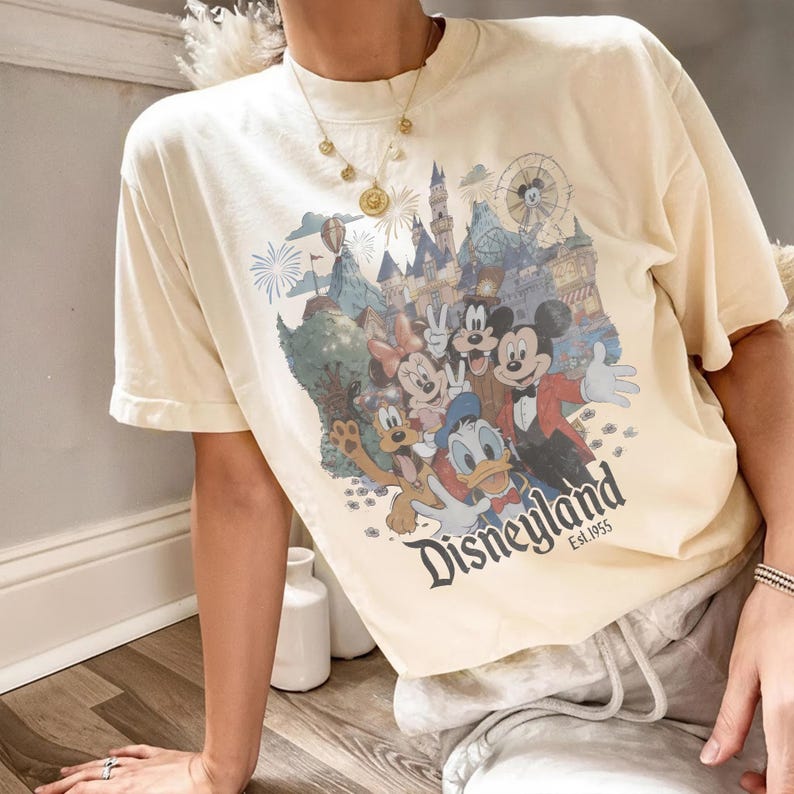 Puede incluir: Camiseta color crema con un gr&aacute;fico colorido de personajes y lugares emblem&aacute;ticos de Disneyland. El gr&aacute;fico incluye a Mickey Mouse, Minnie Mouse, Donald Duck y Goofy. El texto "Disneyland Est. 1955" est&aacute; impreso debajo.