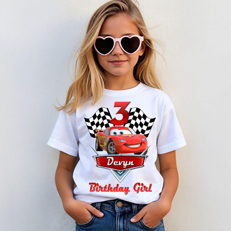 May include: White t-shirt featuring a red cartoon race car, the number 3, and checkered flags. The shirt also has the name "Devyn" and the text "Birthday Girl" in red. The girl is wearing heart-shaped sunglasses.