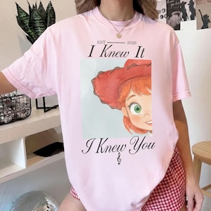 K&ouml;nnte beinhalten: Ein hellrosa T-Shirt mit kurzen &Auml;rmeln und einer Grafik einer Zeichentrickfigur mit roten Haaren und einem roten Cowboyhut. Der Text lautet EST 2026 I Knew It I Knew You, erg&auml;nzt durch ein kleines Notensymbol unter dem Schriftzug.