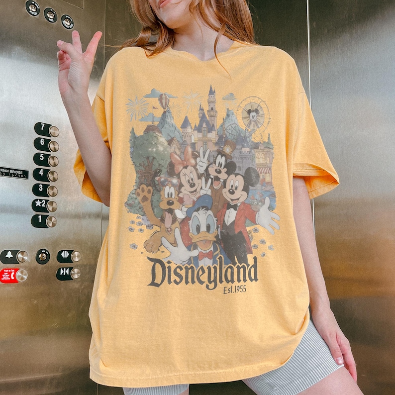 Puede incluir: Camiseta amarilla mostaza con un dise&ntilde;o de Disneyland que presenta a Mickey Mouse, Minnie Mouse, Donald, Goofy y Pluto. El dise&ntilde;o incluye un castillo, fuegos artificiales y el texto "Disneyland Est. 1955".