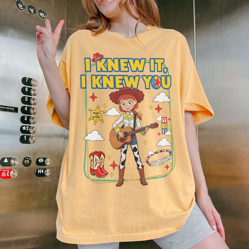 May include: A yellow t-shirt featuring a graphic of a cowgirl character playing an acoustic guitar. The text reads I KNEW IT, I KNEW YOU. Additional graphics include a sheriff badge, cowboy boots, alphabet blocks, and a beaded bracelet.