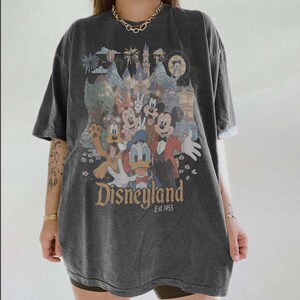 Puede incluir: Camiseta gris oscuro con un gr&aacute;fico colorido de personajes de Disney, incluyendo Mickey Mouse, Minnie Mouse, Donald Duck y Goofy, frente a un castillo. La palabra "Disneyland" est&aacute; impresa en una fuente vintage, con "Est. 1955" debajo.