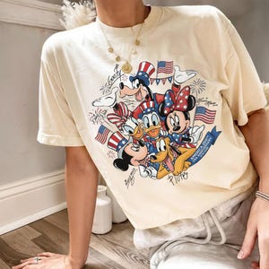 May include: Cream-coloured t-shirt with a graphic of Disney characters in patriotic outfits, including Mickey and Minnie Mouse, Donald Duck, and Goofy. The design includes American flags and the text "America 250th USA Anniversary 1776-2026".