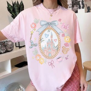 May include: A pale pink t-shirt with a fairytale-themed graphic. The design includes a castle in an oval frame, surrounded by floral and decorative elements. The shirt's colour is a soft, pastel pink.