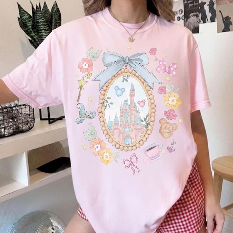 May include: A pale pink t-shirt with a fairytale-themed graphic. The design includes a castle in an oval frame, surrounded by floral and decorative elements. The shirt's colour is a soft, pastel pink.