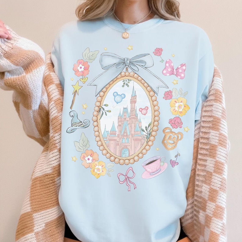 May include: Pale blue sweatshirt with a whimsical design. The design includes an oval frame with a castle, surrounded by flowers, bows, and other decorative elements. A light blue bow is at the top of the frame. The sweatshirt has a crew neck.