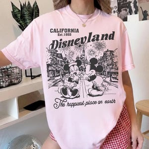 May include: A light pink t-shirt featuring a black and white graphic of Mickey and Minnie Mouse in front of a Disneyland scene. The text "CALIFORNIA Est. 1955 Disneyland" and "The happiest place on earth" are also printed on the shirt.