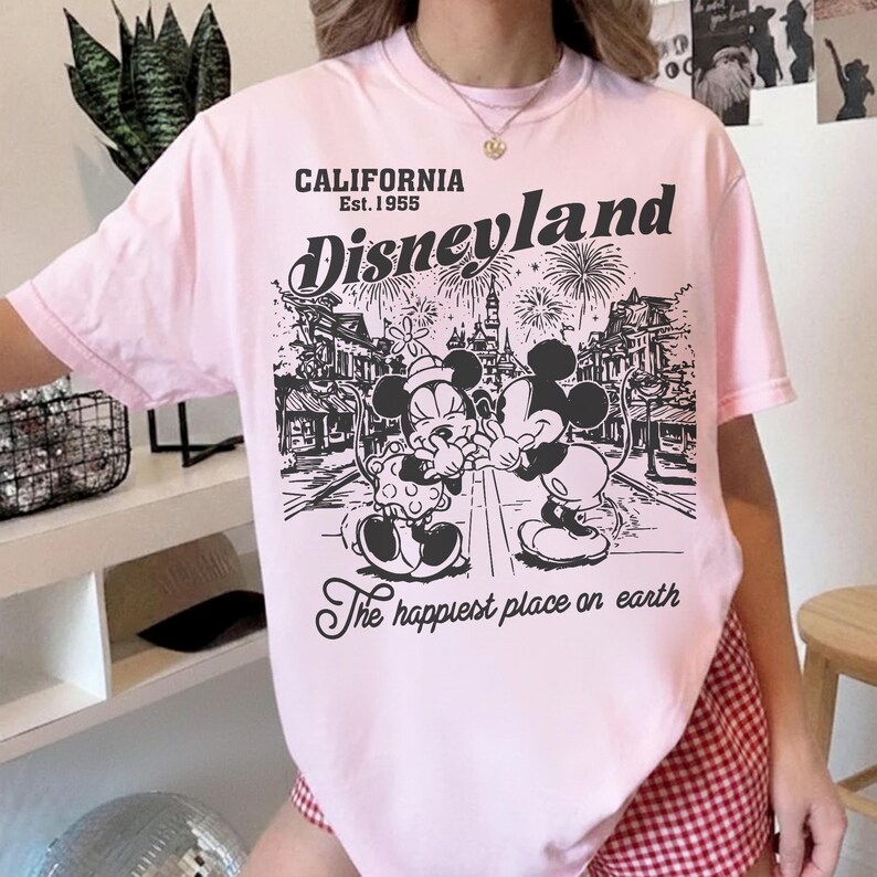 May include: A light pink t-shirt featuring a black and white graphic of Mickey and Minnie Mouse in front of a Disneyland scene. The text "CALIFORNIA Est. 1955 Disneyland" and "The happiest place on earth" are also printed on the shirt.