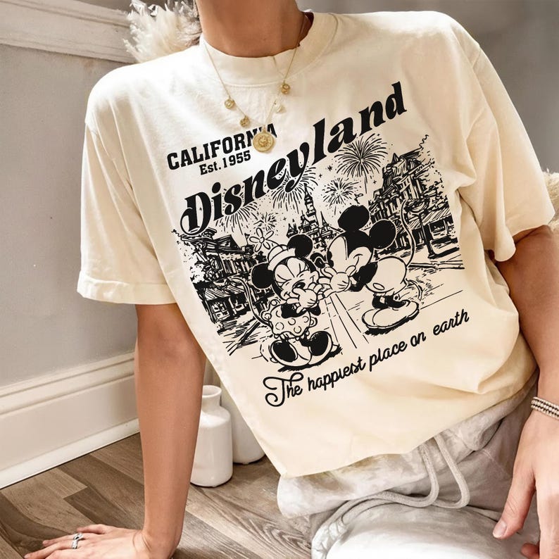 May include: Cream-colored t-shirt featuring a black and white graphic of Mickey and Minnie Mouse in front of a Disneyland scene. The shirt has the text "Disneyland California Est. 1955" and "The happiest place on earth."