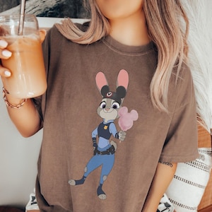 Puede incluir: Camiseta marr&oacute;n con una imagen de dibujos animados de Judy Hopps de Zootopia, vestida de polic&iacute;a y sosteniendo un algod&oacute;n de az&uacute;car rosa. El dise&ntilde;o est&aacute; centrado en la camiseta.