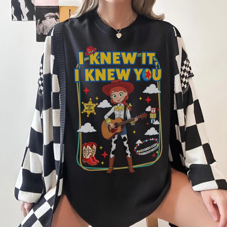 May include: A black t-shirt featuring a graphic of a cowgirl playing a guitar, surrounded by icons like a sheriff star, cowboy boots, and blocks. The text reads I KNEW IT, I KNEW YOU. The shirt is worn under a black and white checkered cardigan.