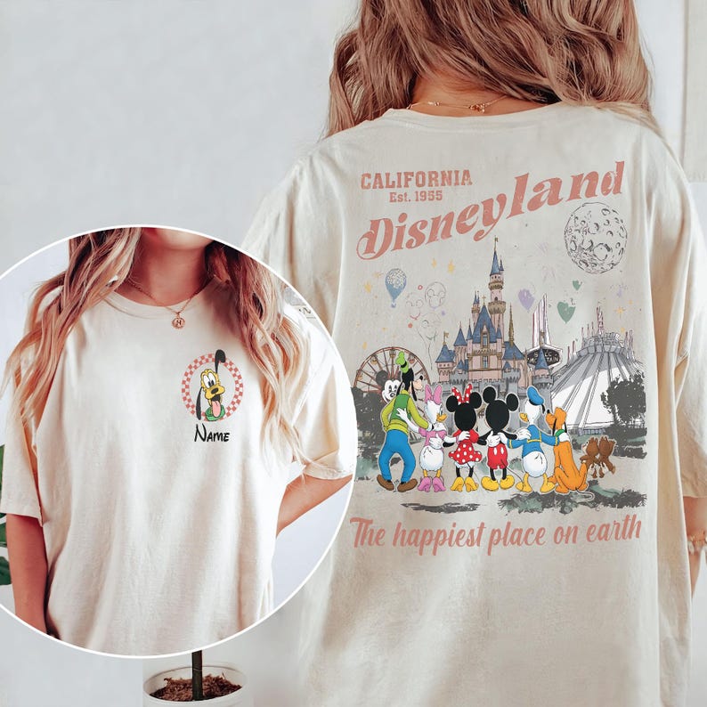 K&ouml;nnte beinhalten: Cremefarbenes T-Shirt mit Disneyland-Design. Auf der Vorderseite ist ein Cartoon von Pluto mit dem Wort "Name" darunter abgebildet. Auf der R&uuml;ckseite sind das Disneyland-Logo, das Schloss und die Charaktere mit dem Text "The happiest place on earth" zu sehen.