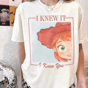 以下が含まれることがあります： クリーム色の半袖Tシャツ。赤い髪と赤いカウボーイハットをかぶった漫画のキャラクターの長方形のグラフィックプリントが施されています。グラフィックの上には「I KNEW IT」、下には「I Knew You」という赤い文字がプリントされています。