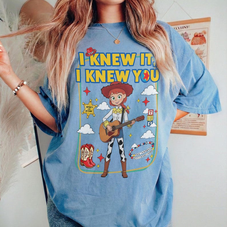 May include: A light blue t-shirt featuring a graphic of a cowgirl character playing an acoustic guitar. The text reads I KNEW IT, I KNEW YOU. The design includes a cowboy hat, a sheriff star, red cowboy boots, clouds, and a beaded bracelet.