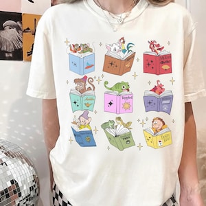 May include: Cream-colored t-shirt featuring colorful illustrations of Disney characters reading books. The books are open, displaying characters from various Disney movies. The design includes stars and the names of the movies.