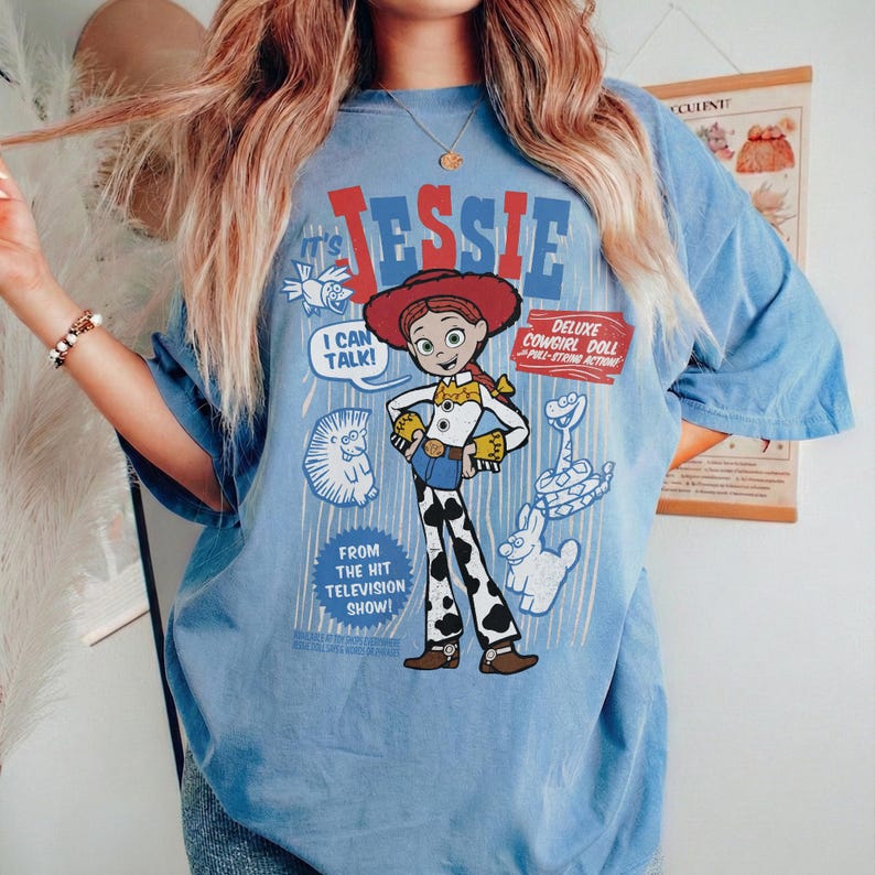 May include: Light blue t-shirt featuring a graphic of the character Jessie from the Toy Story franchise. The design includes the name "Jessie" in red and blue, along with the text "I Can Talk!" and "Deluxe Cowgirl Doll". The shirt also has the text "From the Hit Television Show!"