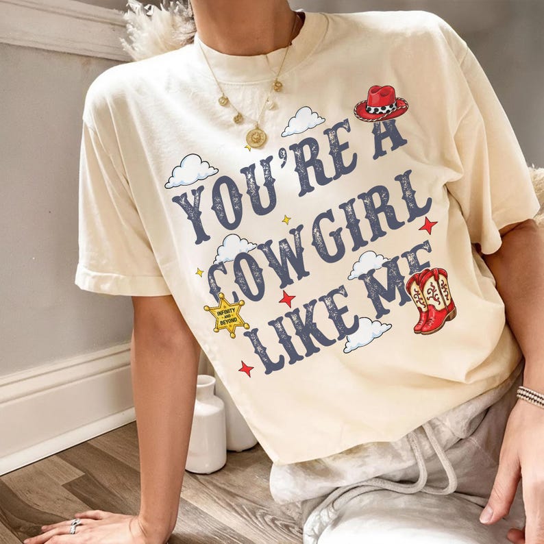 May include: A cream-colored short-sleeve t-shirt featuring the dark blue text "YOU'RE A COWGIRL LIKE ME" surrounded by illustrations of white clouds, red stars, a red cowboy hat, a yellow sheriff badge, and a pair of red and white cowboy boots.