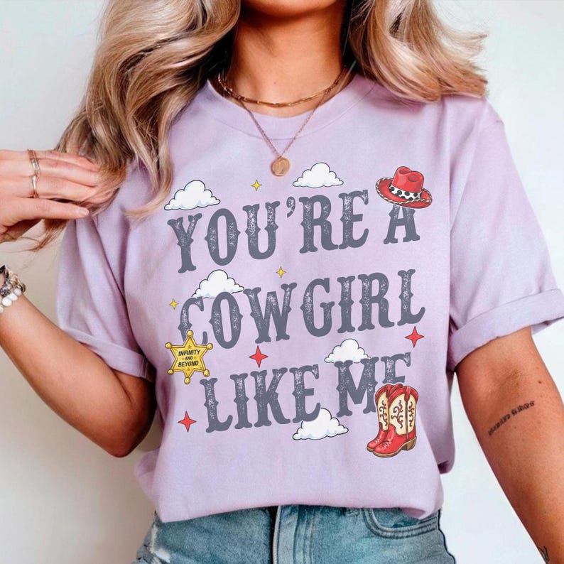 May include: A light purple t-shirt featuring the text "YOU'RE A COWGIRL LIKE ME" in a dark gray western-style font. The design includes small white clouds, red four-pointed stars, a yellow sheriff badge with "INFINITY AND BEYOND" text, and a red cowboy boot.