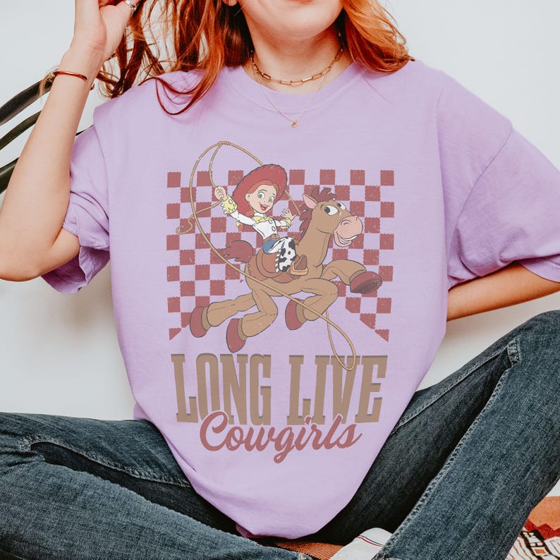 K&ouml;nnte beinhalten: Helllila T-Shirt mit einem Cartoon-Motiv einer Cowgirl auf einem Pferd, vor einem karierten Hintergrund. Der Text "LONG LIVE Cowgirls" ist darunter gedruckt. Das Design ist in Braun- und Rott&ouml;nen gehalten.