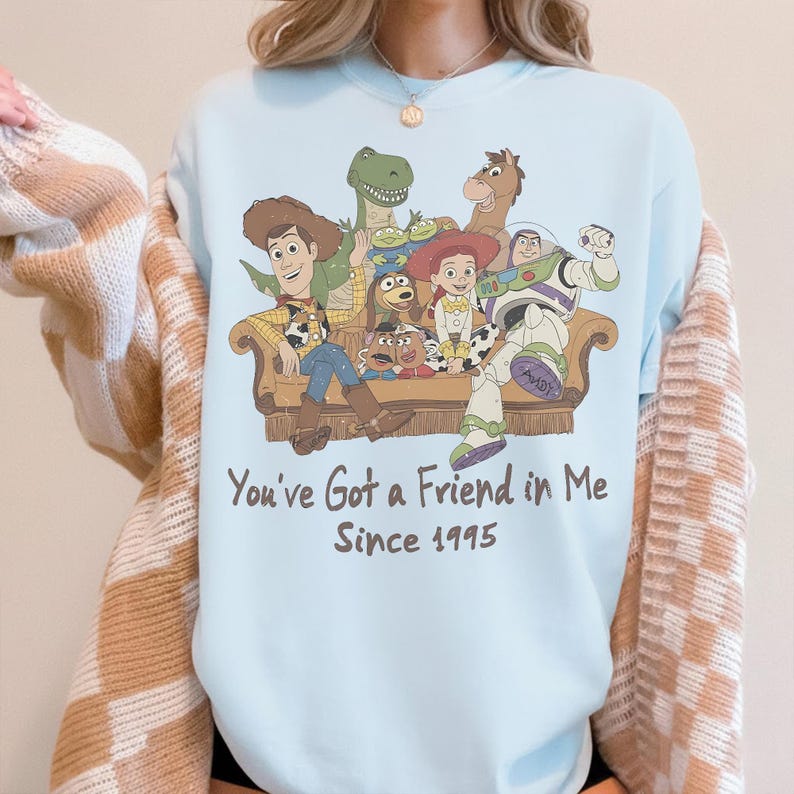 May include: A light blue crewneck sweatshirt featuring a graphic print of Toy Story characters sitting on a couch. Below the illustration, the text reads: You've Got a Friend in Me Since 1995. The sweatshirt is layered under a beige and white checkered cardigan.