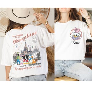 K&ouml;nnte beinhalten: Wei&szlig;e T-Shirts mit Disney-Figuren. Ein Shirt hat eine Disneyland-Grafik auf der R&uuml;ckseite mit dem Text "California Est. 1955" und "The happiest place on earth". Das andere Shirt hat eine Daisy Duck-Grafik mit dem Text "Name".