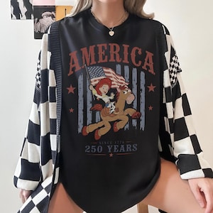 May include: A black crewneck t-shirt featuring a graphic print of a cowgirl riding a horse while holding an American flag. The text reads AMERICA at the top and SINCE 1776 250 YEARS at the bottom. The shirt is worn under a black and white checkered cardigan.