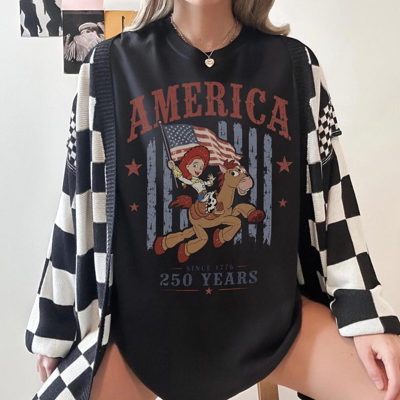 May include: A black crewneck t-shirt featuring a graphic print of a cowgirl riding a horse while holding an American flag. The text reads AMERICA at the top and SINCE 1776 250 YEARS at the bottom. The shirt is worn under a black and white checkered cardigan.
