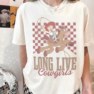 K&ouml;nnte beinhalten: Cremefarbenes T-Shirt mit einem Cartoon-Cowgirl auf einem Pferd, vor einem rot-wei&szlig; karierten Hintergrund. Der Text "LONG LIVE Cowgirls" ist darunter gedruckt. Ein lustiges, l&auml;ssiges T-Shirt.