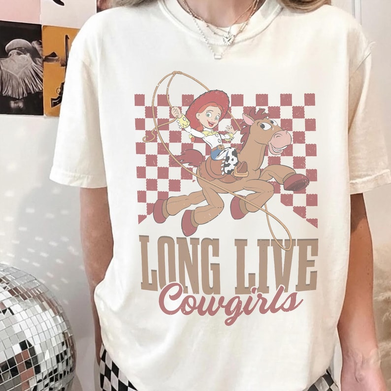K&ouml;nnte beinhalten: Cremefarbenes T-Shirt mit einem Cartoon-Cowgirl auf einem Pferd, vor einem rot-wei&szlig; karierten Hintergrund. Der Text "LONG LIVE Cowgirls" ist darunter gedruckt. Ein lustiges, l&auml;ssiges T-Shirt.