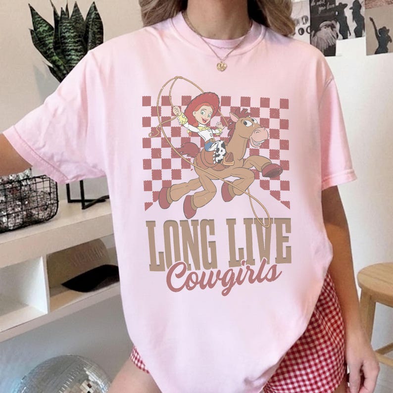 K&ouml;nnte beinhalten: Hellrosa T-Shirt mit einem Cartoon-Cowgirl auf einem Pferd mit Lasso vor kariertem Hintergrund. Der Text "LONG LIVE Cowgirls" ist in Braun und Rosa gedruckt.