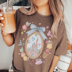 May include: Brown t-shirt with a fairytale-themed design. The design includes a castle, flowers, and a bow. The shirt features a pink teacup, a magic wand, and a butterfly. It's a comfortable, casual style.