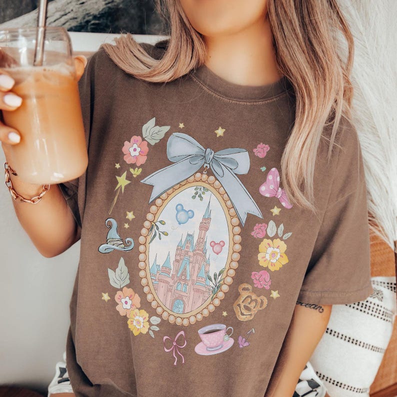 May include: Brown t-shirt with a fairytale-themed design. The design includes a castle, flowers, and a bow. The shirt features a pink teacup, a magic wand, and a butterfly. It's a comfortable, casual style.
