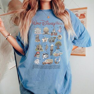 K&ouml;nnte beinhalten: Hellblaues T-Shirt mit einer Grafik von Walt Disney World Attraktionen. Das Design umfasst Illustrationen des Schlosses, Spaceship Earth und anderer ikonischer Wahrzeichen. Der Text "Walt Disney World" ist oben in Rot gebogen.