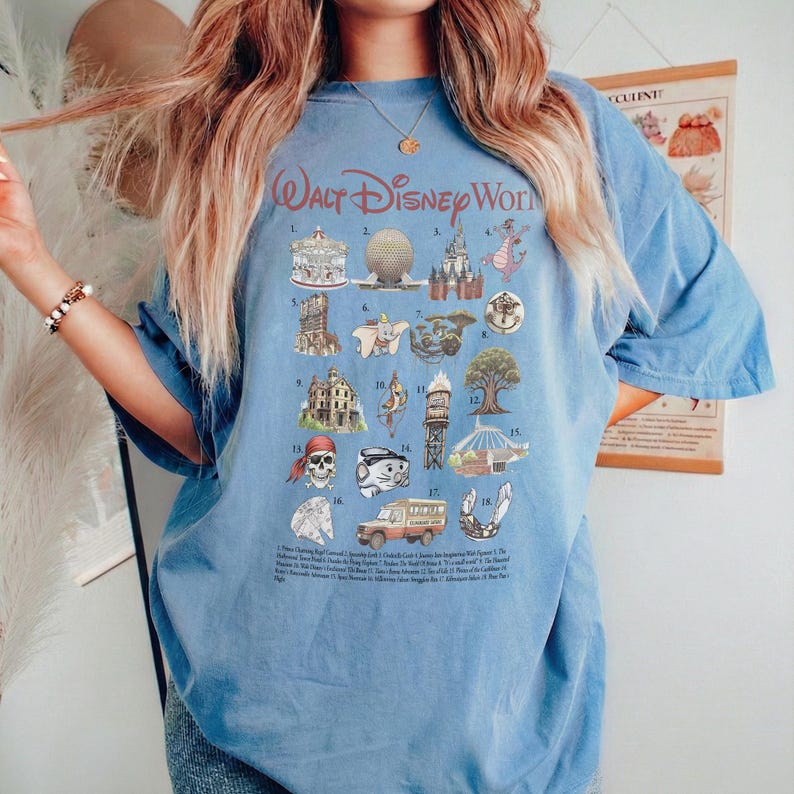 K&ouml;nnte beinhalten: Hellblaues T-Shirt mit einer Grafik von Walt Disney World Attraktionen. Das Design umfasst Illustrationen des Schlosses, Spaceship Earth und anderer ikonischer Wahrzeichen. Der Text "Walt Disney World" ist oben in Rot gebogen.