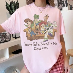 May include: A light pink t-shirt featuring a graphic of Toy Story characters sitting on a couch. The text below reads, "You've Got a Friend in Me Since 1995." The characters include Woody, Buzz Lightyear, Jessie, Rex, Bullseye, Slinky Dog, and Mr. Potato Head.